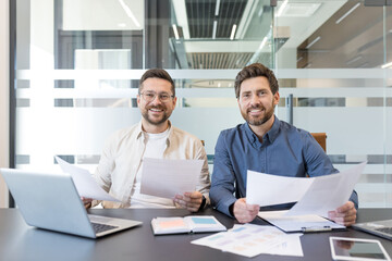 Two smiling colleagues review documents and a laptop at a modern office desk, collaborating on reports and strategy during a productive, friendly business meeting
