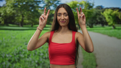 Woman holding air quotes with both hands, showing bare shoulders and midriff while standing on a park path in forest; playful skepticism.