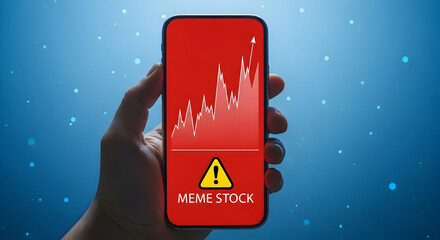 Hand holding smartphone displaying a meme stock chart with a rising trend line and a warning sign on a red screen against a blue background indicating potential investment risk and market volatility