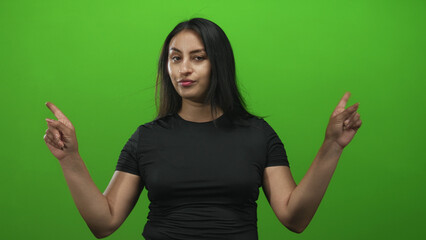 Woman in black shirt points both index fingers upward and holds palms open while shrugging in green studio; doubt indecision.