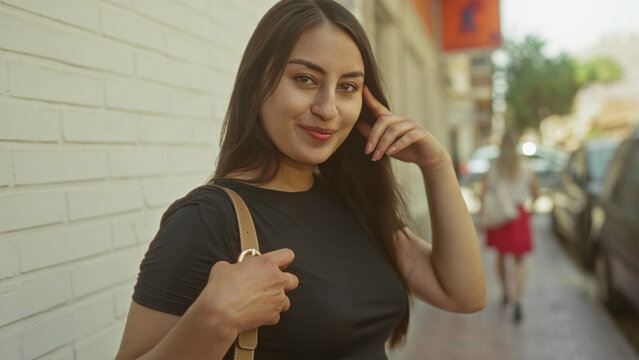 Young smiling woman holding purse strap and touching cheek on sunlit city street; confidence approachability urban charm.
