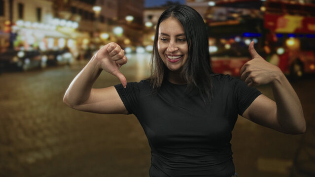 Woman smiling while showing thumbs up and thumbs down with bare thumbs and forearms on a city street at night; ambivalence irony. - Powered by Adobe