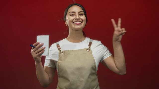 Young woman waitress wearing beige apron holding notepad and pen and showing peace sign while smiling in red studio; cheerful service. - Powered by Adobe