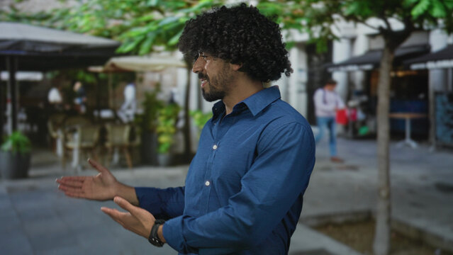 Man with curly hair showing open palms in a street cafe setting while smiling and wearing a blue shirt, presenting the scene; friendly greeting.