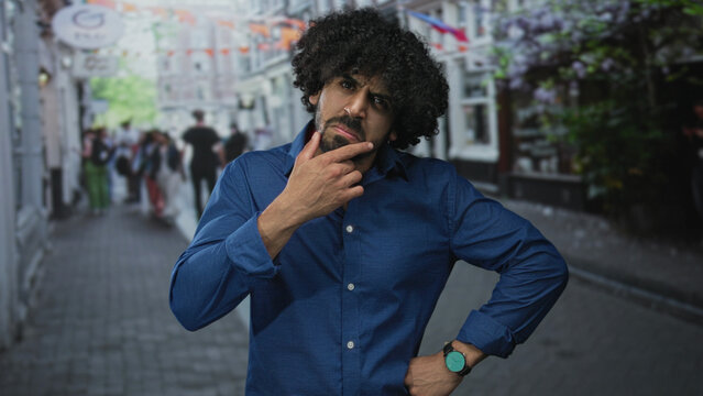 Man with hand on chin on street, curly hair and beard, gesturing with free hand while pausing to think; thoughtful doubt decision reflection. - Powered by Adobe