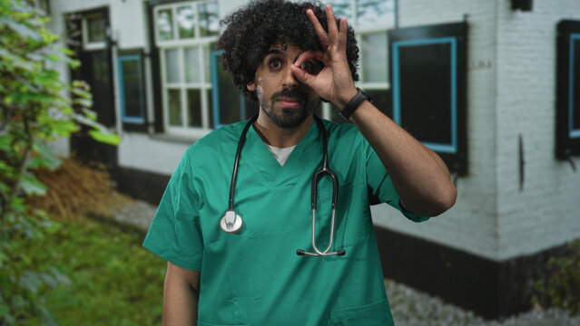 Man doctor in green scrubs with stethoscope making ok sign over eye at building entrance; playful compassion.