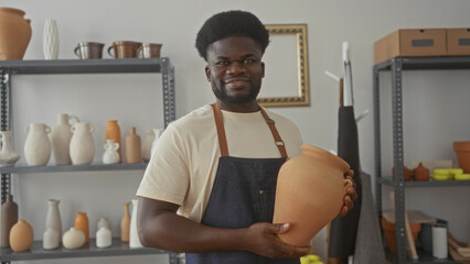 Man potter holding large clay pot in studio with shelves of ceramic vases and wearing apron;...