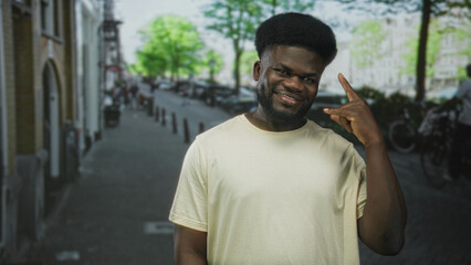 Fototapeta premium Young african american man points finger to camera on city street, smiling and making rock hand gesture while wearing light beige t shirt; confidence connection.