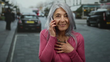 Senior woman with grey hair in a pink sweater talks on smartphone on a bustling street with cars and pedestrians in the background, showcasing urban outdoor communication.