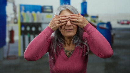 Fototapeta premium Senior woman with grey hair smiling playfully outdoors at a petrol station, spontaneously expressing joy while covering and uncovering her eyes joyfully.