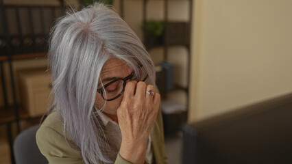Senior woman in office rubbing her eyes with glasses, showing exhaustion in a workplace setting, surrounded by shelves and using a computer in a modern indoor environment.