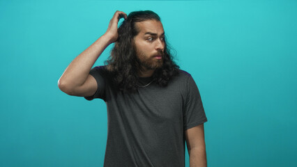 Naklejka premium Man scratching head with hand in studio turquoise backdrop showing beard and long hair; puzzled uncertainty.