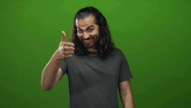 Young arab hispanic man showing thumbs up with visible thumbs and grin in green studio; approval endorsement success. - Powered by Adobe