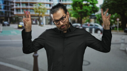 Naklejka premium Man making air quotes with both hands and wearing glasses on a busy city street with buildings and parked cars; playful irony.