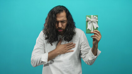 Young man with long curly dark hair and closed eyes holding a green gift and hand on chest in turquoise studio; regret.