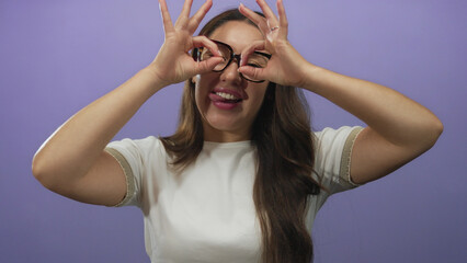 Woman making hand binoculars gesture around eyes and laughing while smiling in purple studio; playfulness curiosity discovery imagination.