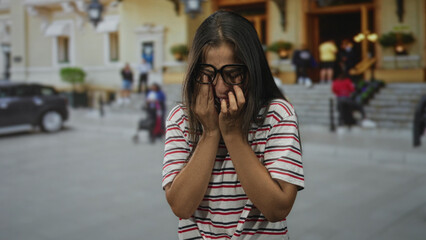 Young woman wearing glasses holding hands to face crying on a city street near building steps;...