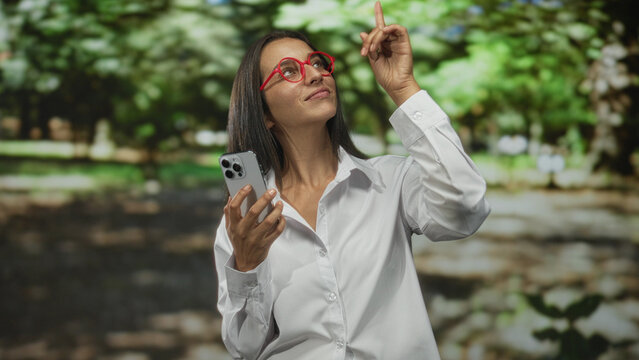 Woman holding smartphone points index finger up in park among green trees and sunlit walkway, smiling while talking on phone; joy connection.