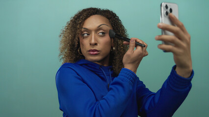 Woman brushes cheek with makeup brush while holding smartphone in studio; confidence selfcare empowerment identity.