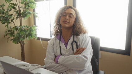Woman doctor in white coat with stethoscope types on laptop while seated at desk in clinic; focus determination perseverance resolve.