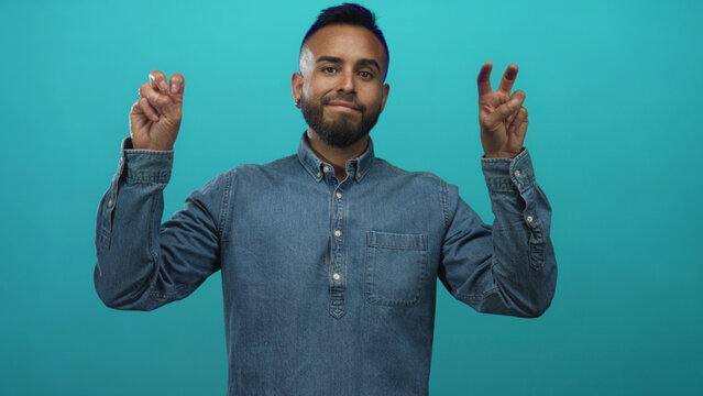 Man in denim shirt making peace sign with both hands in teal studio, facing camera with a slight smile; youthful peaceful optimism.