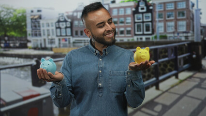 Man smiling holds blue and yellow piggy banks in both bare hands on street by canal bridge in...