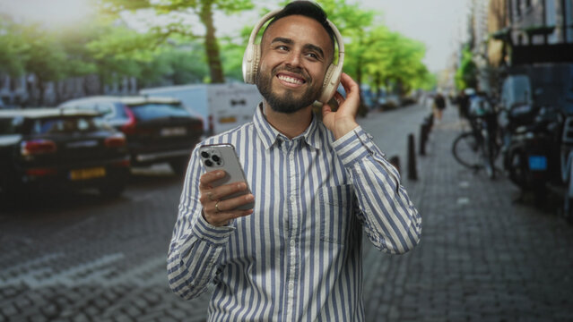 Young hispanic man smiling, holding smartphone in one hand and touching headphones while standing on a cobblestone street; music joy escape.