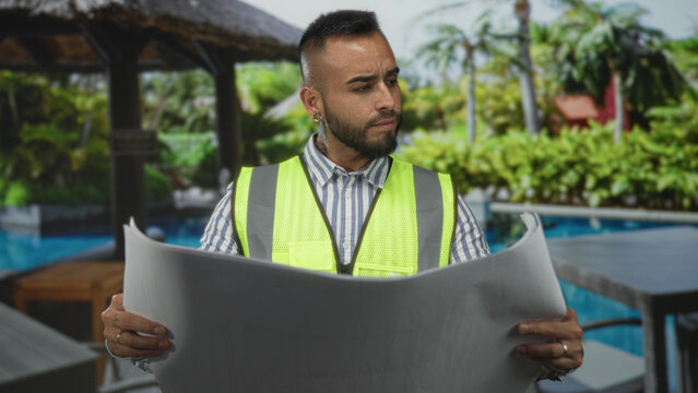 Man holding large blueprint with both hands wearing high visibility vest and striped shirt beside hotel pool and thatched cabana building; concentration.