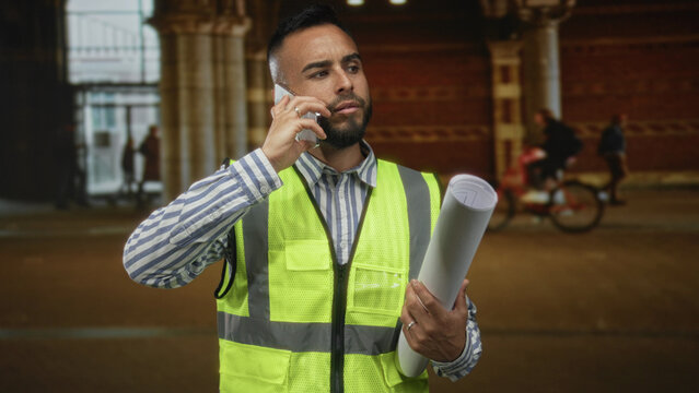 Man engineer holding rolled blueprints and phone to ear while standing in building wearing high visibility vest; professional determination.