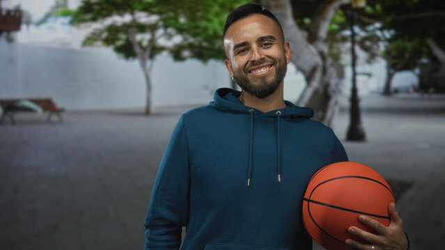 Man in blue hoodie holding a basketball against his hip on a tree lined street plaza, smiling and looking down, tattooed neck and ring on hand; confidence joy. - Powered by Adobe