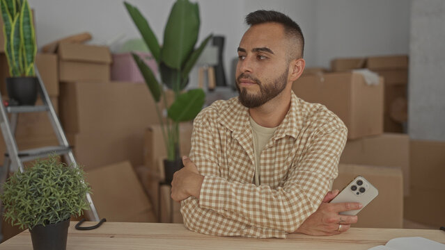 Young hispanic man with arms crossed holding smartphone at wooden table in building filled with moving boxes, ladder and houseplants; moving day contentment.