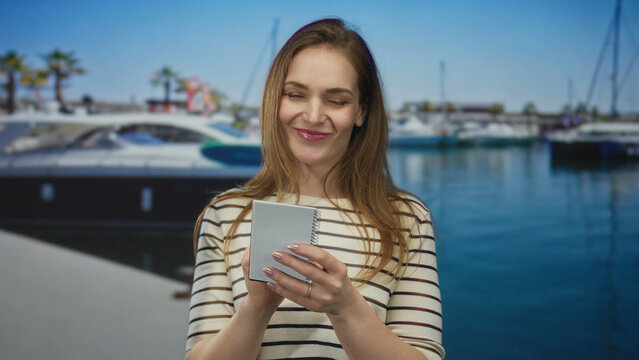 Young woman writing in spiral notebook at sunny port overlooking marina and moored boats; reflection serenity.