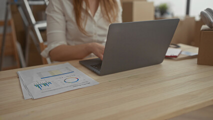 Woman typing on laptop amid unpacked cardboard boxes at wooden table in new home building; focus planning.