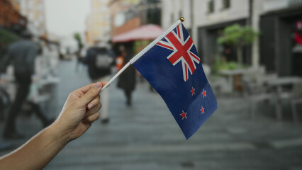 Man holding new zealand flag outdoors on a busy city street with blurred people and buildings in...