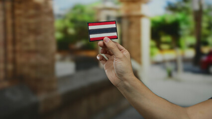 Man holding an embroidered thai flag patch outdoors in a city street setting, showcasing the intricate detail of the fabric design against a blurred urban background.