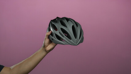 Hand of man holding gray helmet against pink isolated background showcasing safety and cycling gear.