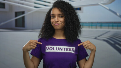 Woman smiling in outdoor setting wearing volunteer shirt, pointing to text while standing with building background and ocean view, showcasing community involvement.
