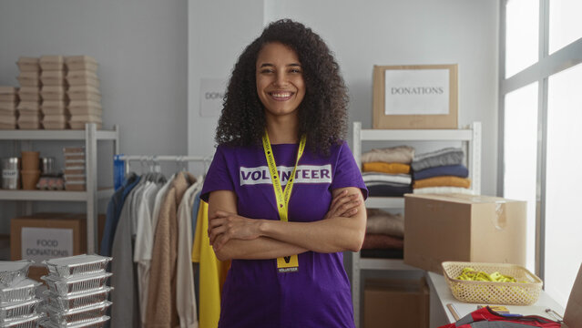 Young hispanic woman crosses arms in indoor volunteer center with donation boxes and clothing shelves; rejection. - Powered by Adobe