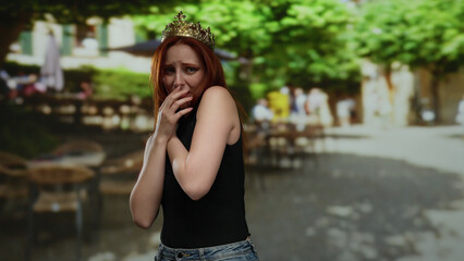 Woman in a crown with red hair stands worried on a bustling city street during daylight.