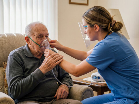 Male senior patient using nebulizer mask with nurse help at home respiratory asthma therapy medical care copd treatment healthcare lungs assistance illness elderly recovery machine inhalation 