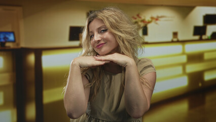 Fototapeta premium Blonde woman resting chin on hands in hotel lobby smiling at camera with soft golden lighting and reception desk visible; confidence.