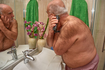 A focused senior citizen cuts his hair and trims his beard in front of the mirror in a simply...