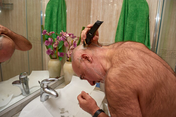 A focused senior citizen cuts his hair and trims his beard in front of the mirror in a simply...