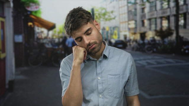Man with eyes closed leaning head on hand on quiet urban street corner lined with old buildings; weariness solitude introspection boredom.