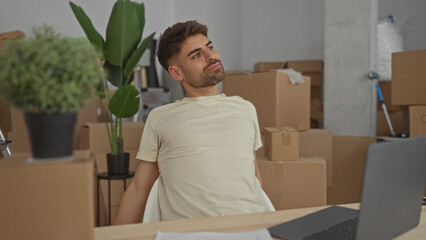 Man stretches neck and arms in a new building with cardboard moving boxes and a laptop on a wooden desk; relief.