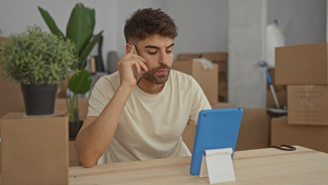 Hispanic man holds smartphone to ear and taps tablet screen among cardboard boxes in building; preparation.