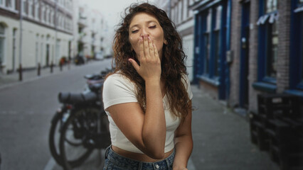 Woman smiling and blowing kiss with hand to lips on street showing midriff and casual jeans; joy carefree warmth.