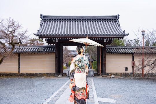 A traditional Japanese young woman with traditional Japanese-style house with dark brown wooden exterior, dark grey tiled roofed gate