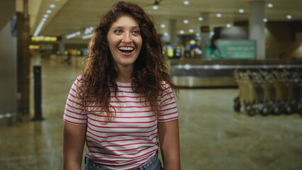 Woman with open mouth smiling, freckled face and bare forearms visible in busy airport terminal near baggage carousels and trolleys; arrival joy.