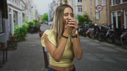 Fototapeta premium Woman laughing with hands covering mouth on a street lined with bikes and buildings, wearing yellow crop top showing navel piercing; joyful spontaneity.